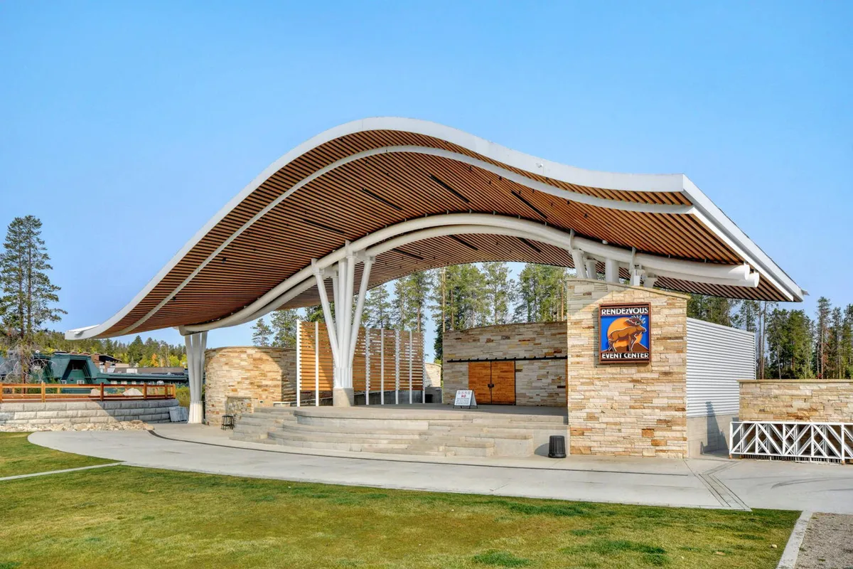 An exterior shot of the Hideaway Park amphitheater, showcasing its distinctive curved roof, stone facade, and wooden elements. The setting is ideal for outdoor concerts, community gatherings, and festivals, with an uplifting and communal feel.