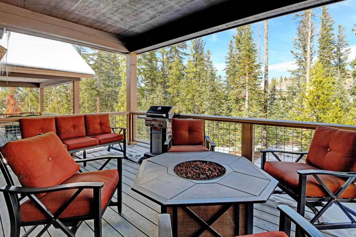 An inviting outdoor deck scene with comfortable seating, a glowing fire pit, and a grill, all nestled among tall trees. The ambiance is warm and welcoming, suggesting cozy evenings socializing by the fire or gathering for a barbecue as the sun sets behind the mountains.