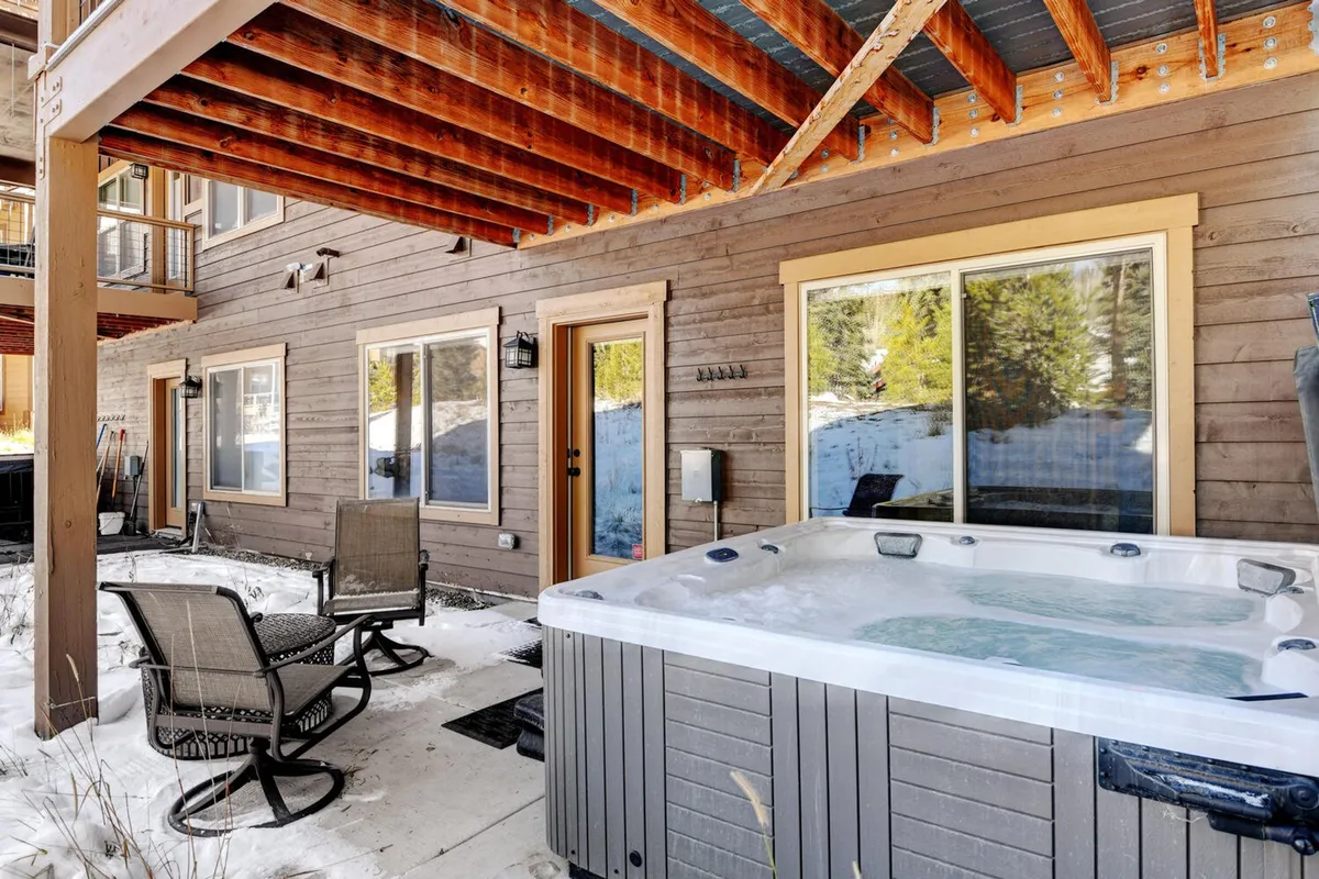 An inviting patio scene features a bubbling hot tub, cozy outdoor seating, and wooden beams, all set against the backdrop of the home’s exterior walls. The setup suggests opportunities for socializing and relaxing after a day spent outdoors, with the hot tub as a focal point.