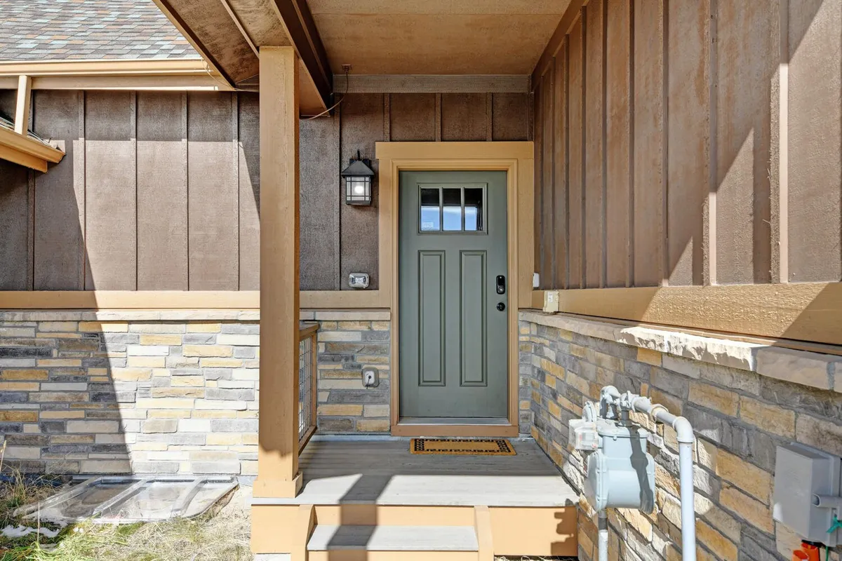 The image shows the exterior front door of the chalet, framed by a stone wall and a warmly lit entrance. A welcome mat and lighting fixture create an inviting first impression, suggesting a cozy and exclusive retreat awaiting inside.