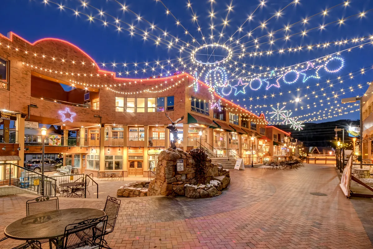 A cheerful nighttime exterior featuring string lights overhead, decorative stars, a stone fountain, and outdoor seating. The scene conveys a festive and romantic atmosphere, ideal for evening strolls, community events, and alfresco dining.