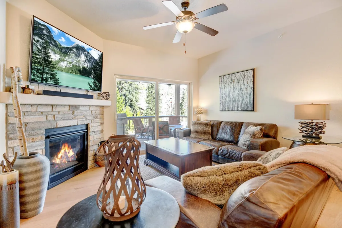 The image showcases a spacious, inviting living room with plush sofas arranged around a glowing stone fireplace and a large TV. Expansive windows offer breathtaking mountain views, while the warm lighting and chic décor create an atmosphere perfect for relaxing, socializing, or taking in the sunset after a day of adventure.