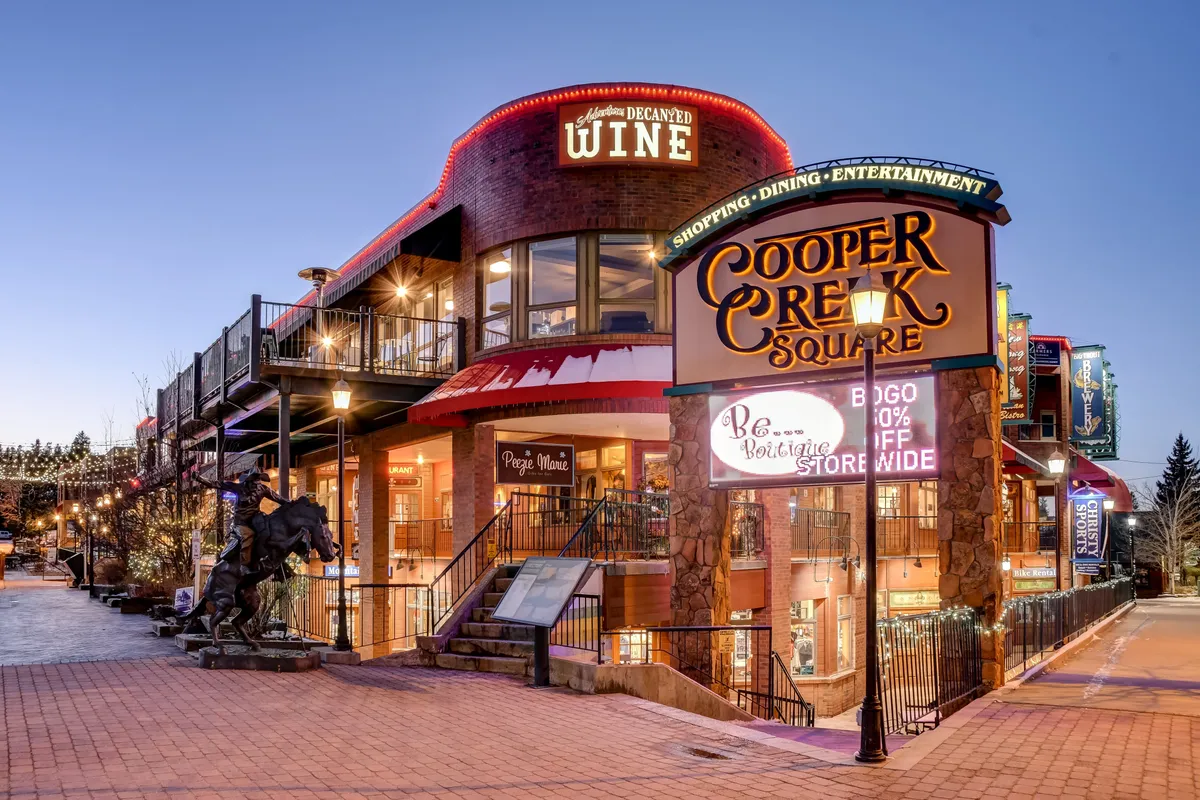 The image features the Cooper Creek Square sign, the entrance to a wine shop, an outdoor seating area, decorative lighting, and a sculpture of a horse. The scene is lively and hints at a bustling, social environment perfect for shopping, dining, and local entertainment.
