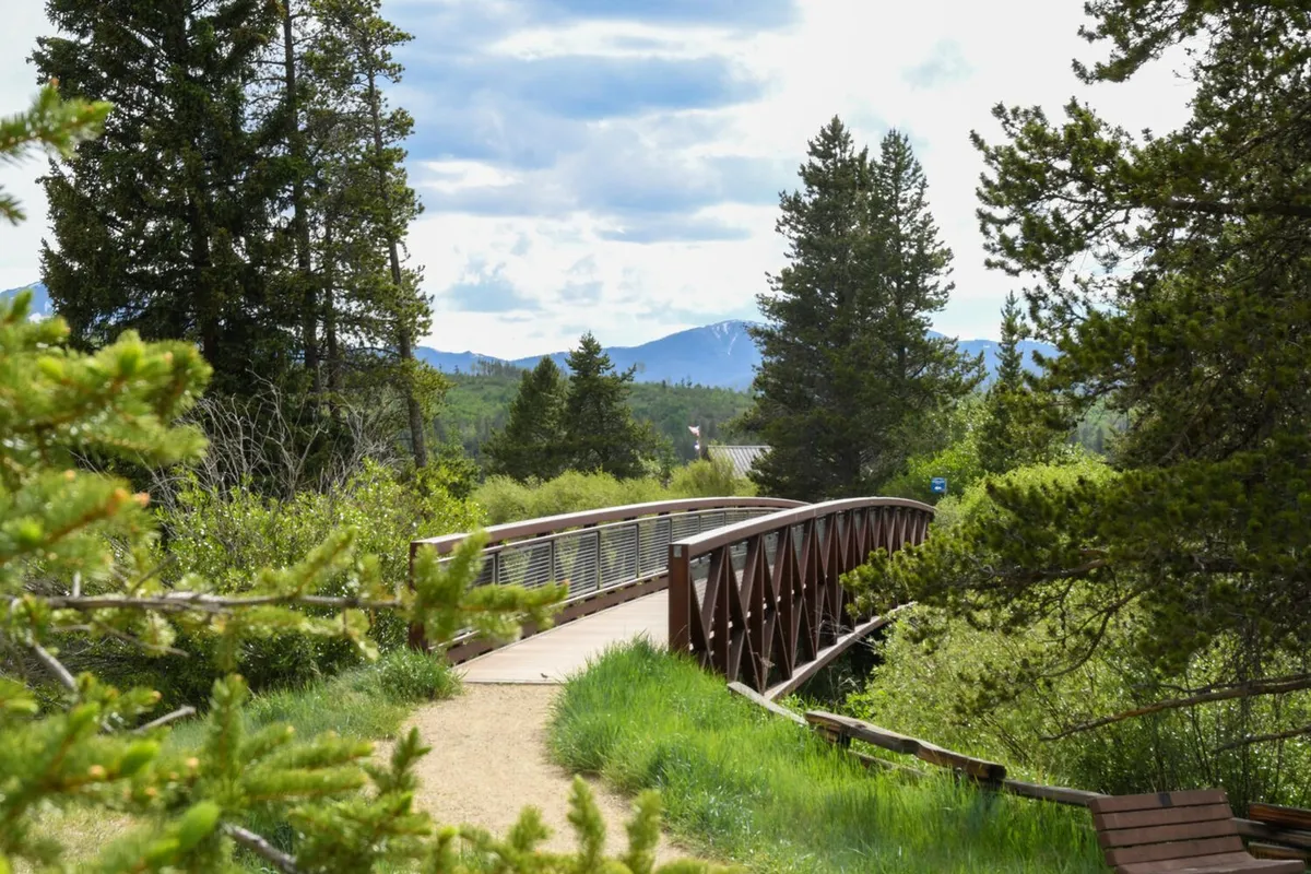 A scenic exterior shot of a wooden bridge crossing through a lush, tree-lined area with a breathtaking mountain backdrop. The tranquil setting is ideal for walking or biking and hints at the extensive trail network accessible from the neighborhood.