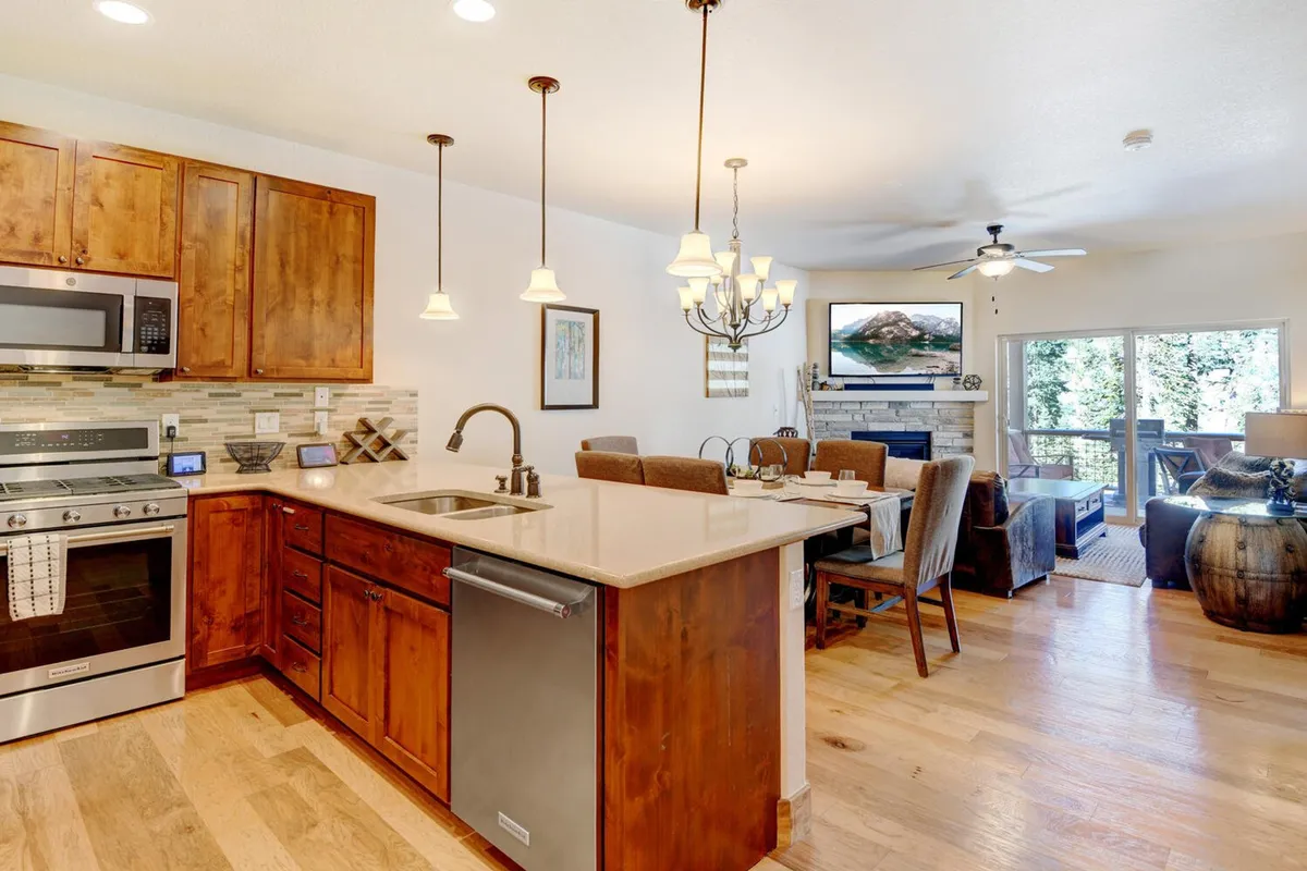 A wide-angle shot encompasses the open-plan living area, visually connecting the kitchen island, dining table, and a comfortable seating area arranged around a glowing fireplace. The space is bathed in natural light from large windows, with subtle mountain vistas visible in the background.