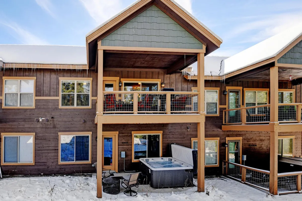 This exterior shot features a spacious balcony with a hot tub, wooden siding, and large windows that look out onto winter scenery. The inviting hot tub sits at the edge of the balcony, suggesting a relaxing retreat after time outdoors. The setting is tranquil, with hints of Rocky Mountain views visible beyond the deck.